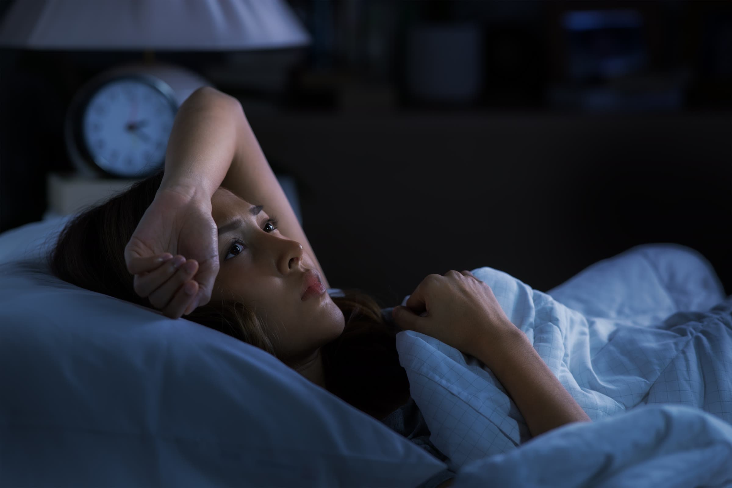 A woman lying awake in bed at night, one hand on her forehead, with a blurred alarm clock on the bedside table.
